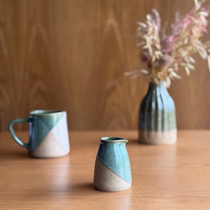 May include: A ceramic creamer, mug, and vase in teal and beige. The creamer has a spout and a diagonal teal section. The mug has a handle and a similar teal and beige design. The vase is green and beige and holds dried flowers.