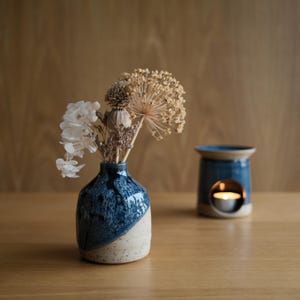 May include: A small ceramic vase with a speckled cream base and a glossy blue glazed top, containing a bouquet of dried flowers including white hydrangea and allium. Beside it sits a matching blue ceramic oil burner with a lit tea light candle.