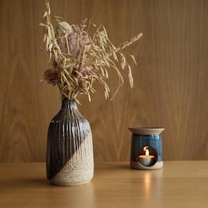 May include: A ceramic vase with a ribbed texture, featuring a dark brown glazed upper section and a speckled cream base, filled with dried grasses and flowers. Beside it sits a blue ceramic oil burner with a lit tea light candle inside.