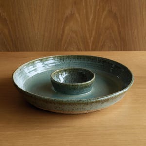 May include: A round ceramic serving dish with a central integrated bowl, featuring a mottled sage green and brown glaze with a glossy finish. The wide outer rim surrounds the smaller inner bowl, creating a unified piece with a textured base.