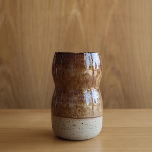 May include: A cylindrical ceramic vase with a pinched middle section. The upper portion features a glossy, mottled brown glaze with reflective highlights, while the lower base is composed of a matte, off-white clay with dark speckles throughout.