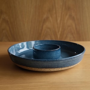 May include: A round ceramic serving dish with a deep blue speckled glaze and a central integrated bowl. The exterior base of the dish features a contrasting unglazed, light beige speckled texture, while the interior has a smooth blue finish.