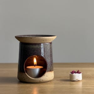 May include: A dark brown ceramic oil diffuser with a speckled tan rim and base, with a lit tea light candle inside. A small white wax melt with dried flower petals sits to the right. The diffuser is on a wooden surface.