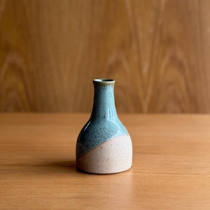 May include: A small ceramic bud vase with a narrow neck and a rounded base. The upper portion features a glossy, speckled teal glaze, while the lower section is finished in a matte, speckled beige clay, meeting in a diagonal line across the body.