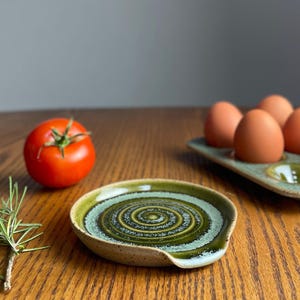 May include: A green and white ceramic spoon rest with a spiral design. The spoon rest is on a wooden table.