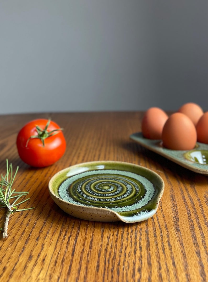 May include: A green and white ceramic spoon rest with a spiral design. The spoon rest is on a wooden table.
