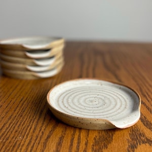 May include: A white ceramic spoon rest with a speckled glaze and a swirl design. The rest is sitting on a wooden surface.