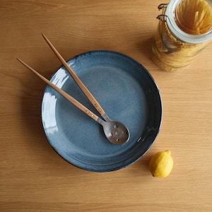 May include: A round, blue ceramic plate with a mottled finish, featuring two long-handled serving utensils with wooden grips and metal heads, one of which is perforated. A clear glass jar of pasta and a single yellow lemon are nearby.