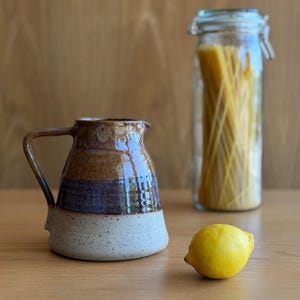 May include: A ceramic pitcher with a glossy finish featuring horizontal bands of brown, dark purple, and speckled cream. Beside it sits a single yellow lemon and a tall glass jar filled with long pasta noodles, all arranged on a wooden surface.