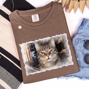 May include: Espresso-colored t-shirt featuring a cat with a lit cigarette. The cat image is framed with a torn paper effect. The shirt is laid out with a tote bag, a necklace, and denim shorts.
