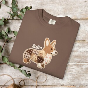 May include: A brown t-shirt featuring a patchwork corgi design with the name "Stella" above it. The corgi is made of various patterned fabrics, including plaid, floral, and polka dots. The shirt is displayed on a wooden surface.