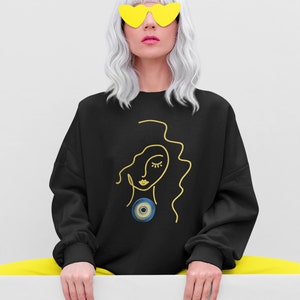 May include: Black sweatshirt with a gold line drawing of a woman's face with a blue and gold evil eye pendant. The model is wearing yellow heart-shaped sunglasses and yellow pants.