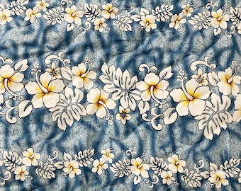 Op de afbeelding: Een blauwe stof met een wit en geel bloemenpatroon. Het patroon toont hibiscusbloemen met gele centra en witte bloemblaadjes. De stof heeft een gestikte textuur.