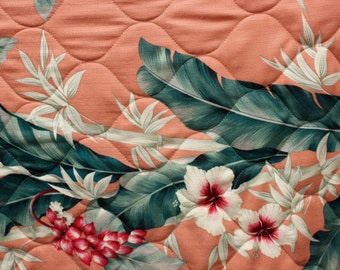 Op de afbeelding: Een gestikte stof met een tropische print met grote groene bladeren, witte bloemen met rode centra en een lichtroze achtergrond.