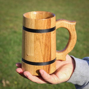 May include: A handcrafted wooden beer mug with a large handle. The mug is made of light brown wood with two black bands around the body. The mug is held in a person's hand against a green background.