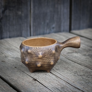 May include: A dark brown wooden cup with a single handle, featuring a rounded body with several protruding, knob-like bumps on the exterior surface. The cup has a visible wood grain pattern and a smooth, circular opening at the top.