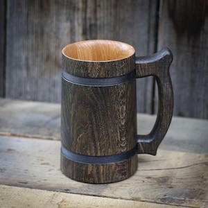May include: A handcrafted wooden beer mug with a dark, stained exterior and a lighter wood interior. The mug features a sturdy handle and is accented with two dark bands. The wood grain is visible, adding to the rustic appeal.