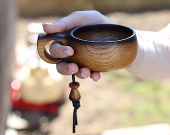 May include: A small, round wooden cup with a single handle featuring a dark brown, gradient finish and visible wood grain. A black cord with a small, matching wooden bead and two dangling ends is attached to the handle of the drinking vessel.
