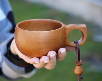 以下が含まれることがあります： 茶色の革ひもが付いた木製マグカップ。マグカップは天然木で作られており、丸みを帯びた形をしています。ひもはマグカップの取っ手に取り付けられており、先端に小さな木製のビーズが付いています。