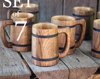 以下が含まれることがあります： 木製のビールジョッキ7個セット。各ジョッキは明るい茶色の木材で作られており、大きなハンドルと中央に暗いバンドが付いています。素朴な木製の表面に配置されています。「SET of 7」の文字が白で表示されています。