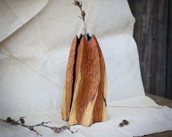 May include: A wooden vase with a unique, twisted design. The vase is made of natural wood with a reddish-brown color and a dark brown stripe running down the center. The vase is sitting on a white fabric surface.