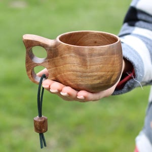 Puede incluir: Una taza de madera con asa y un cordón verde oscuro con una cuenta de madera. La taza está hecha de madera marrón oscura y tiene una forma redonda y lisa.