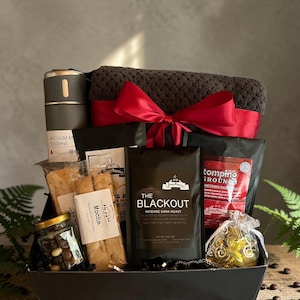 May include: A black gift basket filled with coffee, snacks, and a dark gray blanket tied with a red ribbon. The basket includes a vacuum bottle, coffee beans, and a bag of "The Blackout" intense dark roast coffee.
