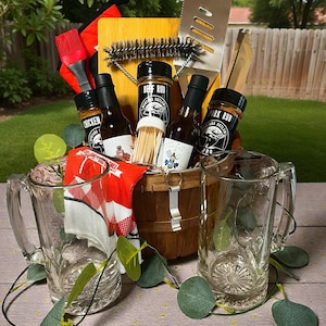 May include: A wooden basket containing two glass beer mugs, a bamboo cutting board, a metal spatula, a grill brush, a red silicone basting brush, a red and white checkered towel, toothpicks, and bottles of Montana Outlaw BBQ chicken, beef, and pork rub.