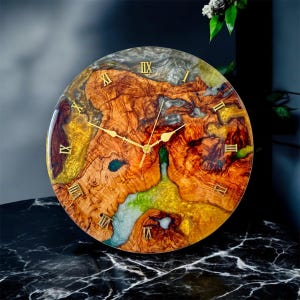 Może przedstawiać: Okrągły zegar ścienny z unikalnym wzorem słojów drewna. Tarcza zegara ma mieszankę brązowych, złotych i zielonych odcieni, ze złotymi rzymskimi cyframi i wskazówkami. Zegar ma średnicę około 30 cm.