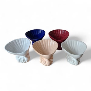 May include: Four ceramic seashell-shaped bowls in different colours: white, pink, blue, and red. The bowls are on a white background.