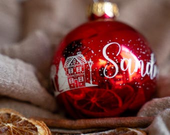 Pode incluir: Um enfeite de Natal esférico vermelho brilhante com um gráfico impresso em branco de uma casa e o nome "Sandra" em letra cursiva. O enfeite tem uma tampa metálica dourada no topo e está rodeado por fatias de laranja seca e canela.