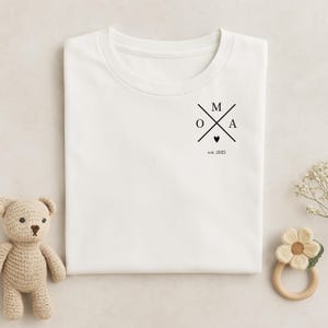 Könnte beinhalten: Weißes T-Shirt mit einem schwarzen Grafikdesign mit den Buchstaben OMA, die mit den Buchstaben M und A gekreuzt sind, einem kleinen Herzen und dem Text "est. 2025". Ein Teddybär und ein Beißring aus Holz liegen neben dem Shirt.