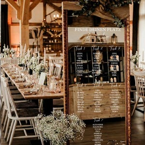 Puede incluir: Un espejo rectangular alto con un marco oscuro y ornamentado que muestra un plano de asientos de boda. El texto dice FINDE DEINEN PLATZ, Anna & Leon, y enumera nombres bajo siete mesas. Está decorado con vegetación y junto a frascos de vidrio.