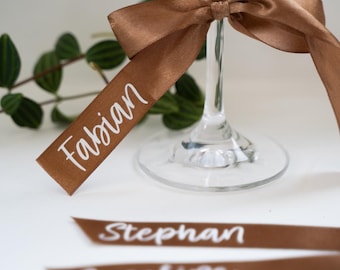 Pode incluir: Uma taça de vinho transparente com uma fita castanha amarrada ao redor da haste. A fita tem o nome "Fabian" impresso em branco. Fitas adicionais com nomes são visíveis.