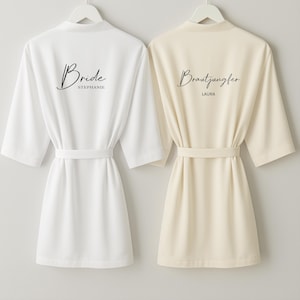 Peut inclure: Deux peignoirs à manches courtes, un blanc et un crème, sont suspendus côte à côte. Le peignoir blanc porte l'inscription "Bride" et un nom au dos. Le peignoir crème porte l'inscription "Bräutigam" et un nom au dos. Chaque peignoir a une ceinture assortie.