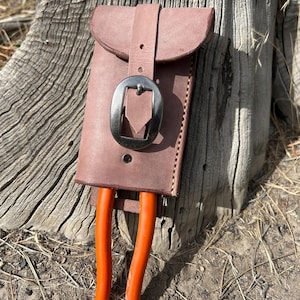 May include: A brown leather tool pouch with a flap and a silver buckle. The pouch holds a pair of orange handled gardening shears. The pouch is attached to a brown leather belt.