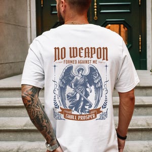 May include: A white t-shirt featuring a graphic on the back with the text NO WEAPON FORMED AGAINST ME SHALL PROSPER and Isaiah 54:17. The design includes a central illustration of an angel with large wings holding a sword, framed by decorative elements.