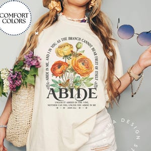 May include: Cream-coloured t-shirt with a floral design featuring yellow and orange flowers and the word "ABIDE." The shirt also includes the text "ABIDE IN ME, AND I IN YOU AS THE BRANCH CANNOT BEAR FRUIT OF ITSELF." The shirt is part of the Comfort Colors collection.