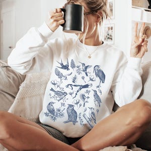 May include: White sweatshirt featuring a blue illustration of various birds and floral elements. The long-sleeved top has a crew neck and is made of a soft material. The sweatshirt is a comfortable and stylish choice for casual wear.