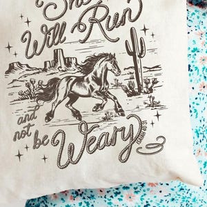 May include: Cream tote bag with a western design. It features a running horse illustration, cacti, and the text "She Will Run and not be Weary." The bag is held by a person.