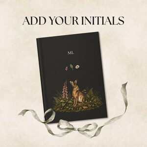 May include: A black notebook with a textured cover featuring a brown rabbit illustration surrounded by green foliage and pink flowers. The initials ML are printed in white above the rabbit. The text ADD YOUR INITIALS appears above the notebook.