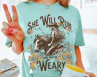 May include: A light green t-shirt with a graphic of a woman riding a horse, surrounded by flowers. The text reads "She Will Run And Not Be Weary" and "Isaiah 40". The shirt has a relaxed fit.