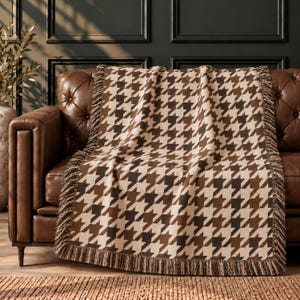 Peut inclure: Un plaid tissé présentant un motif pied-de-poule marron et crème. Ce textile rectangulaire arbore un design géométrique répétitif et est agrémenté de franges décoratives sur les bords, conférant un aspect texturé à l'ensemble du tissu.