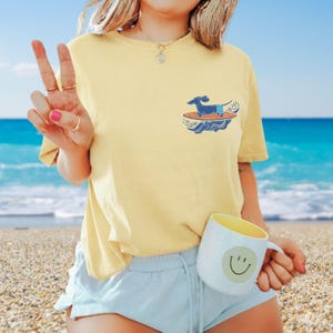 Yellow t-shirt with a small graphic of a dachshund surfing on a blue surfboard. The shirt is paired with light blue shorts. A white mug with a yellow interior and a smiley face is held in the hand.