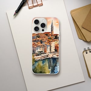 May include: A white phone case featuring a watercolor painting of a coastal town. The artwork depicts buildings with orange rooftops, a tall church tower, and boats on the water. The case has a glossy finish and a protective bumper.