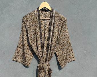 May include: A brown and black patterned robe with a white trim and a black tie belt. The robe is hanging on a wooden hanger against a gray wall.
