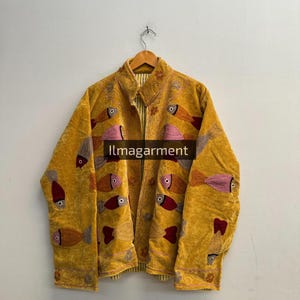 Pode incluir: Casaco amarelo mostarda com design de peixes bordados. O casaco tem gola e fecho de botões. Os peixes são em tons de rosa, laranja e vermelho. O texto "llmagarment" é visível no casaco.