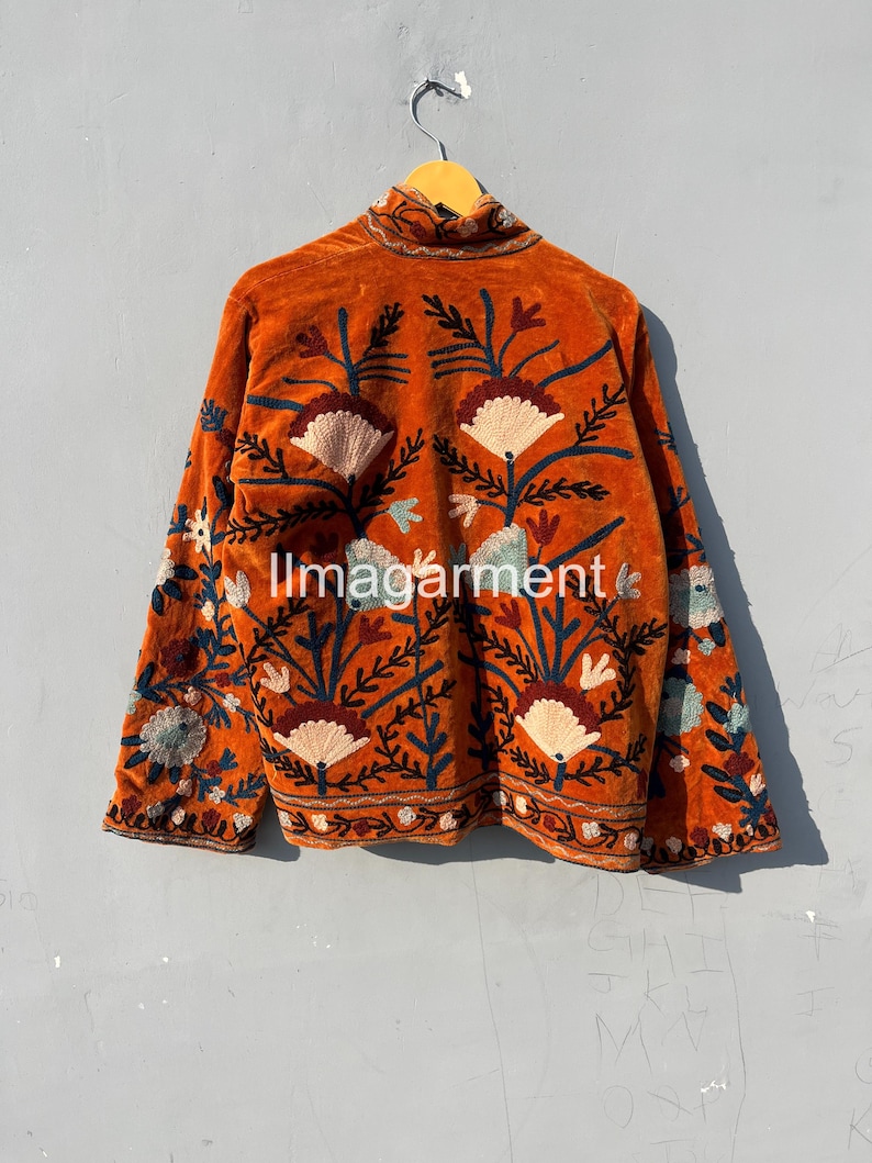 May include: A burnt orange velvet jacket with intricate floral embroidery in shades of blue, green, and pink. The jacket has a high neck and long sleeves.
