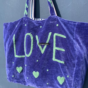 May include: A large, plush, purple tote bag with the word "LOVE" in green, embroidered letters. The bag features green heart and dot accents. The handles are striped with green and white. The bag is made of a soft, velvety material.