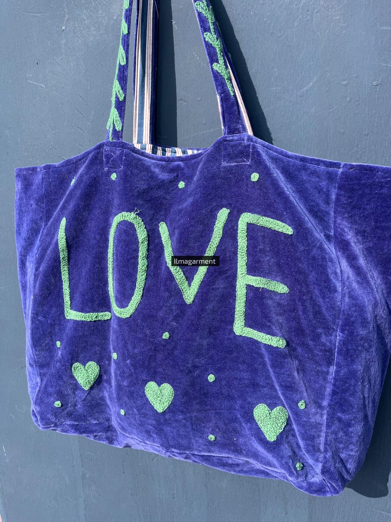 May include: A large, plush, purple tote bag with the word "LOVE" in green, embroidered letters. The bag features green heart and dot accents. The handles are striped with green and white. The bag is made of a soft, velvety material.